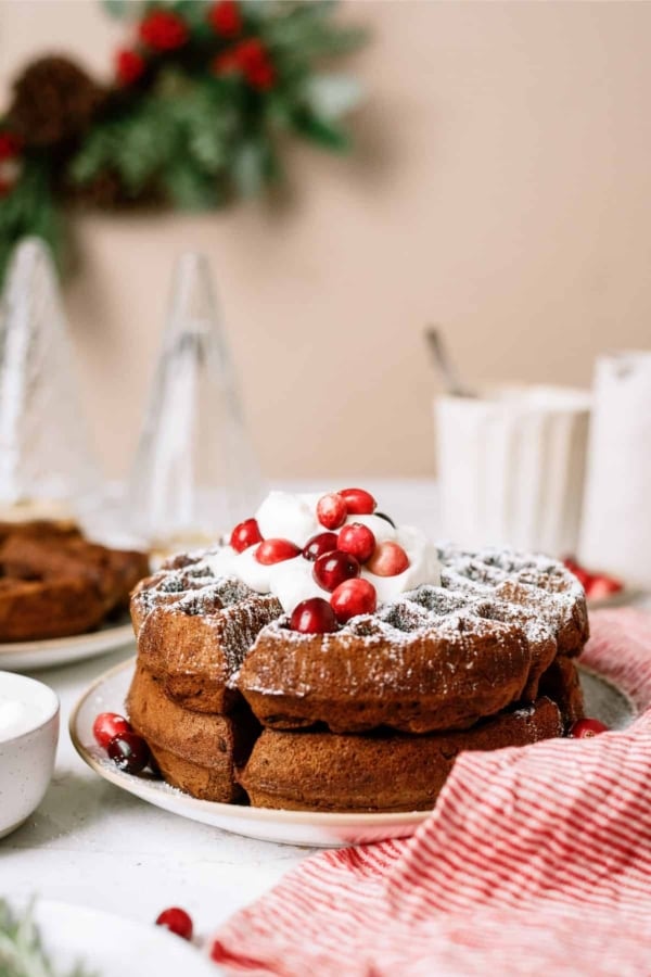 A stack of waffles topped with whipped cream and cranberries on a white plate, with festive decorations and folded red-striped napkin in the background.