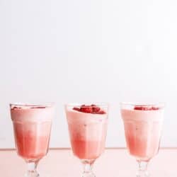 Three glasses filled with pink mousse or dessert, each topped with small pieces of red fruit, are arranged in a row on a light pink surface against a white background.