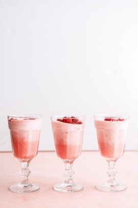 Three glasses filled with pink mousse or dessert, each topped with small pieces of red fruit, are arranged in a row on a light pink surface against a white background.