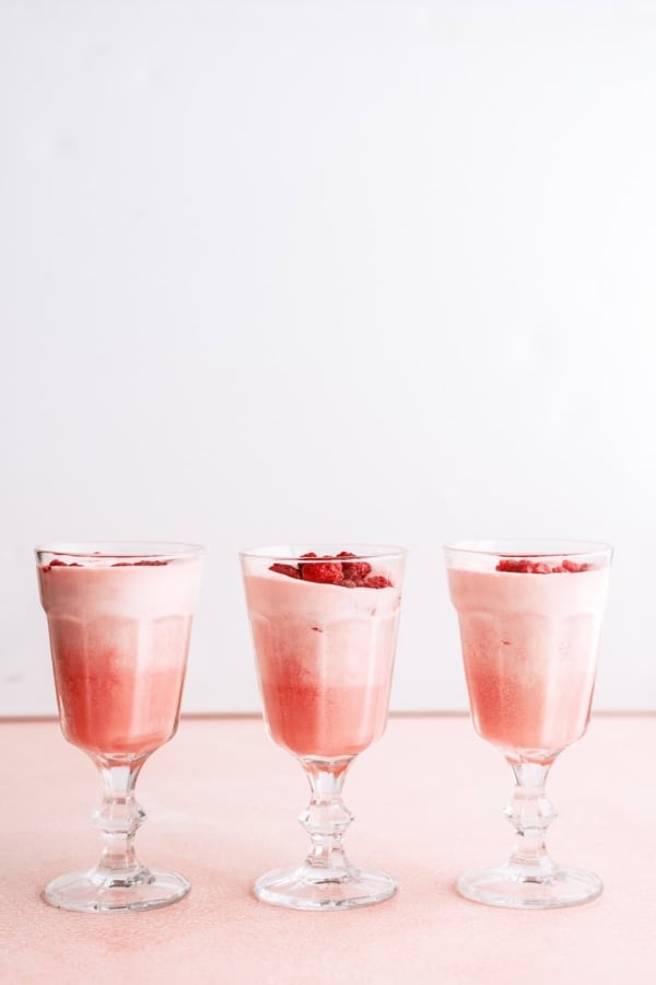 Three glasses filled with pink mousse or dessert, each topped with small pieces of red fruit, are arranged in a row on a light pink surface against a white background.