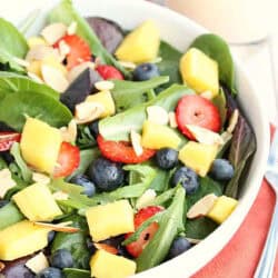 A bowl of mixed green salad topped with pineapple chunks, blueberries, sliced strawberries, and almond slivers, placed on an orange napkin with a fork beside it.