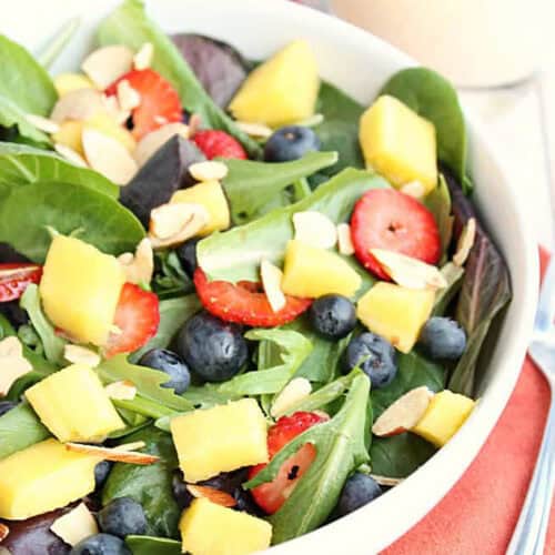 A bowl of mixed green salad topped with pineapple chunks, blueberries, sliced strawberries, and almond slivers, placed on an orange napkin with a fork beside it.