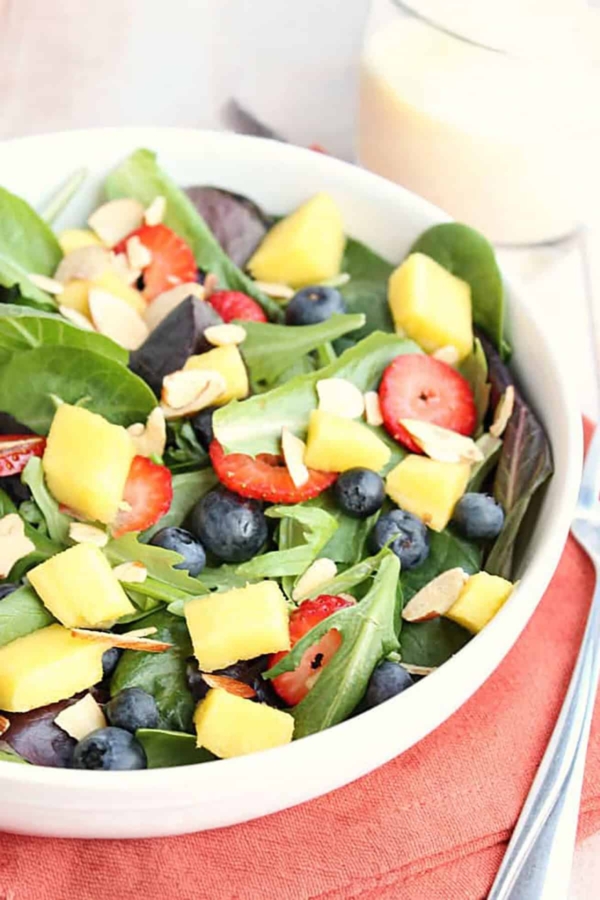 A bowl of mixed green salad topped with pineapple chunks, blueberries, sliced strawberries, and almond slivers, placed on an orange napkin with a fork beside it.