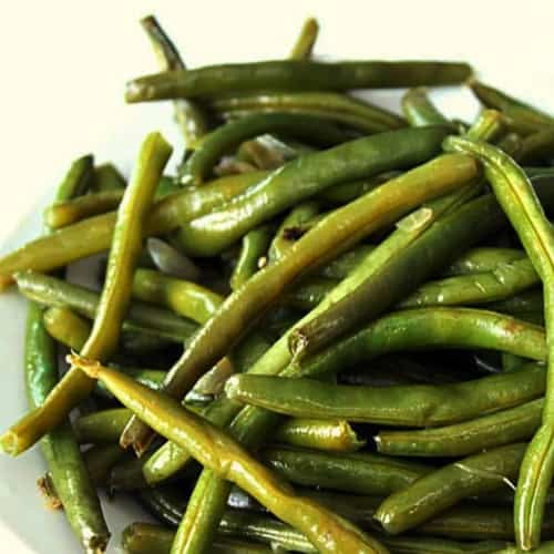 A plate of cooked green beans is arranged in a pile on a white surface.