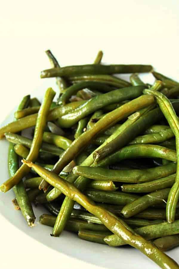 A plate of cooked green beans is arranged in a pile on a white surface.