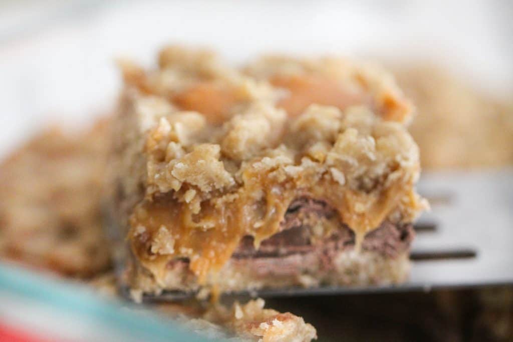Close-up of a fork holding a square piece of dessert featuring layers of chocolate, caramel, and crumble topping.
