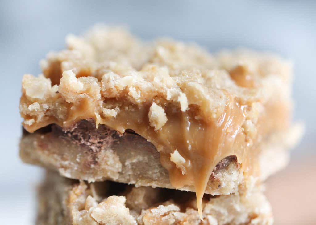 A closeup shot of a stack of three caramel oat bars with gooey caramel and chocolate layers sits on a white plate.