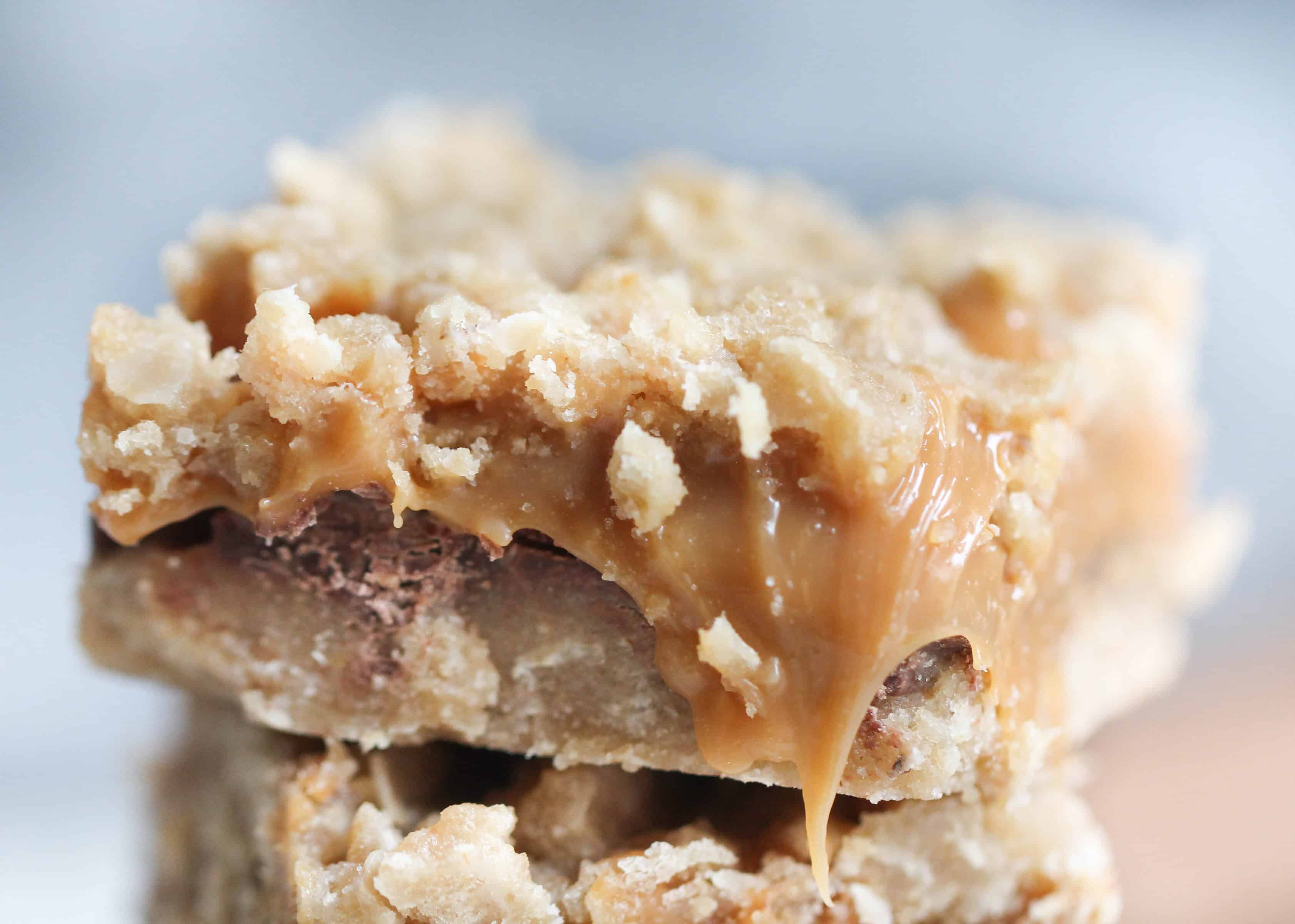 a closeup shot of a stack of three caramel oat bars with gooey caramel and chocolate layers sits on a white plate.