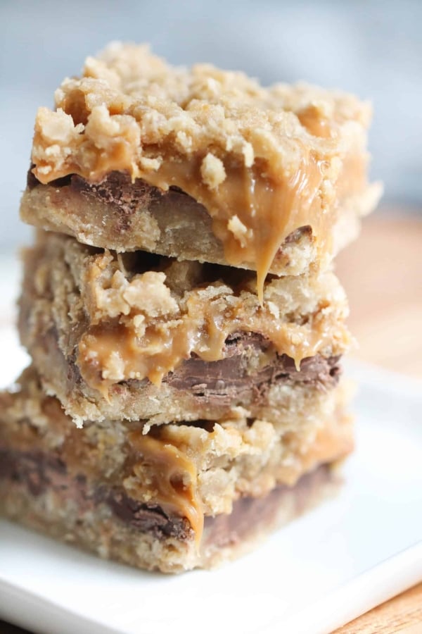 A stack of three caramel oat bars with gooey caramel and chocolate layers sits on a white plate.