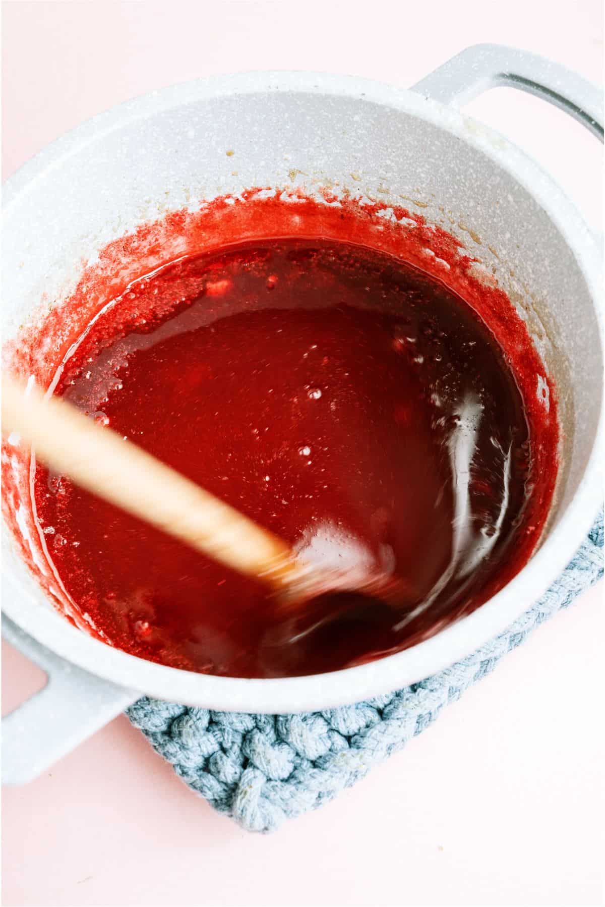 Strawberry jello mixture in a saucepan with a wooden spoon.