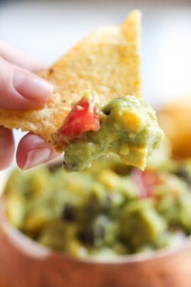 A hand holds a tortilla chip with a scoop of guacamole, containing chunks of tomato and corn. The background shows more guacamole.