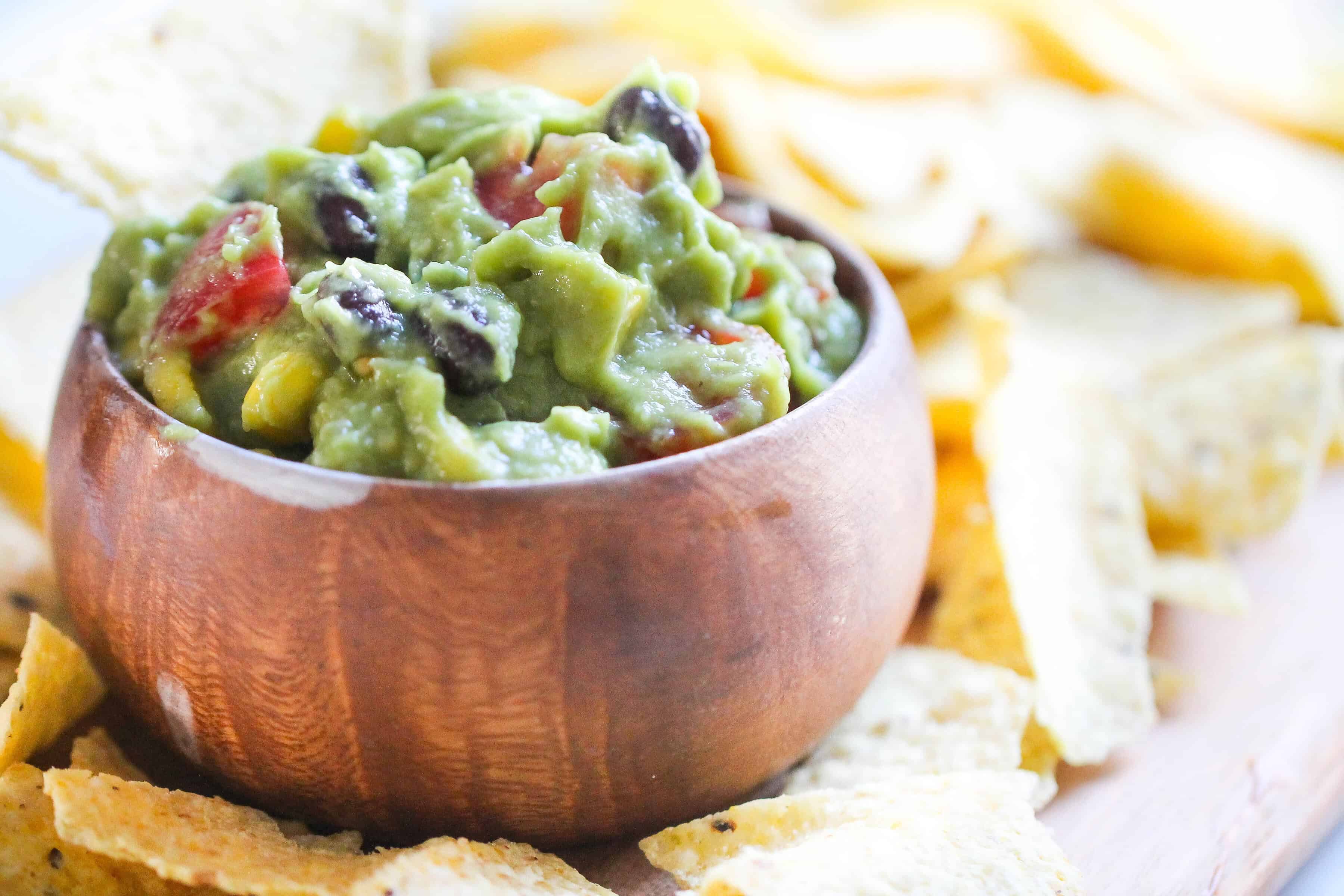 A wooden bowl filled with guacamole, surrounded by tortilla chips. The guacamole contains ingredients like tomatoes, corn, and black beans.
