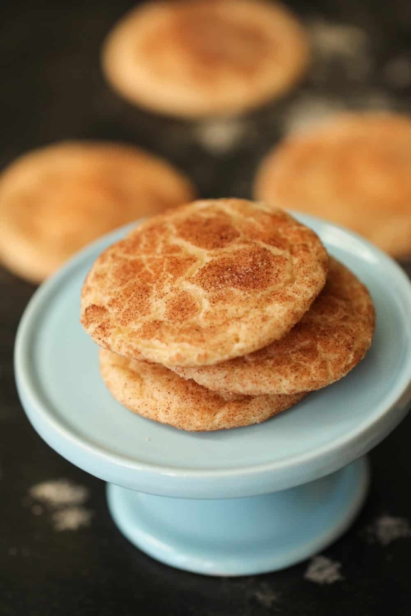 3 Snickerdoodle Cookies on top of a small cake stand with remaining Snickerdoodle Cookies in the background.