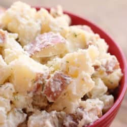 A red bowl filled with creamy potato salad, containing chunks of potatoes, visible seasoning, and potato skins.