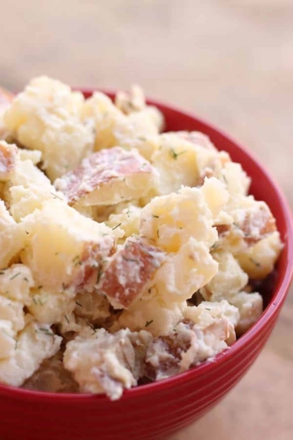 A red bowl filled with creamy potato salad, containing chunks of potatoes, visible seasoning, and potato skins.