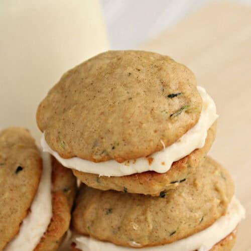 Close-up of three zucchini whoopie pies with cream filling stacked on a light-colored surface with a blurred milk bottle in the background.