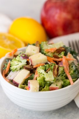 A white bowl filled with broccoli salad mixed with carrots, apple chunks, and nuts. A halved lemon and a red apple are in the background.