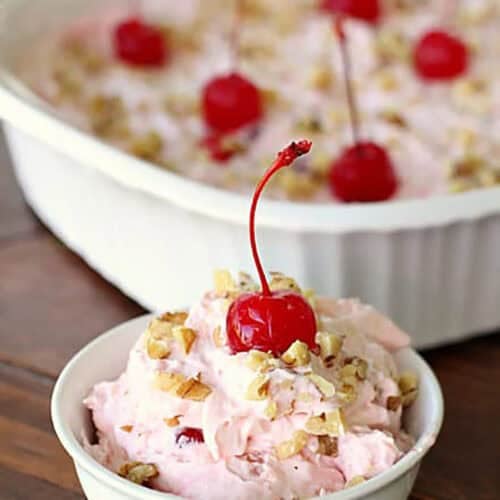 A small bowl of pink dessert topped with a cherry and nuts in front of a larger, matching dish with the same dessert.