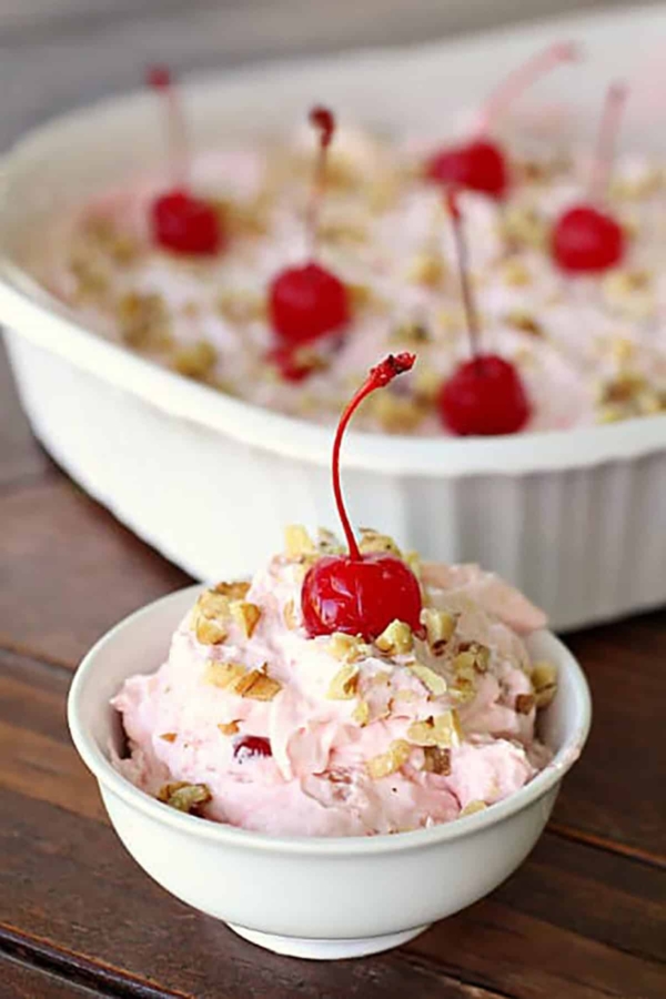A small bowl of pink dessert topped with a cherry and nuts in front of a larger, matching dish with the same dessert.