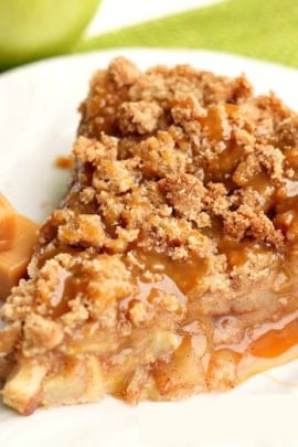 A slice of caramel apple crumble pie on a white plate, topped with caramel sauce and accompanied by caramel candy pieces.
