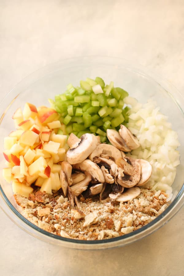A glass bowl containing chopped apples, celery, onions, mushrooms, and breadcrumbs.