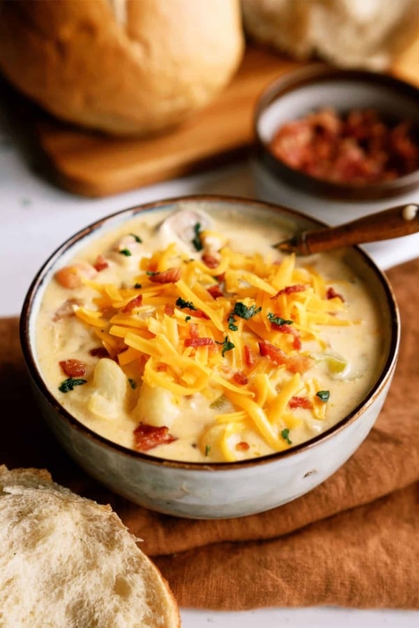 A bowl of creamy soup topped with shredded cheddar cheese, chopped bacon, and herbs, with a slice of bread on the side.