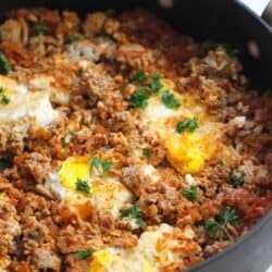 Eggs poached in a tomato-based sauce with ground meat and herbs in a black skillet.