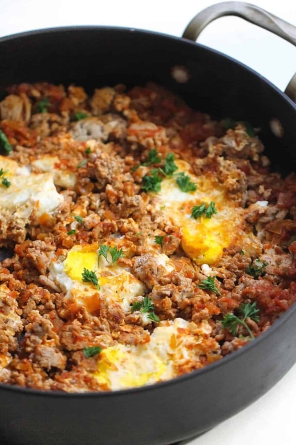 Eggs poached in a tomato-based sauce with ground meat and herbs in a black skillet.
