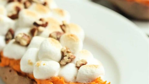A baked sweet potato half topped with toasted marshmallows and chopped pecans is served on a white plate.