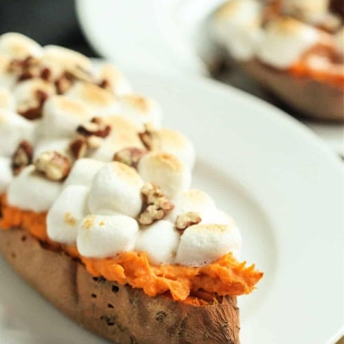 A baked sweet potato half topped with toasted marshmallows and chopped pecans is served on a white plate.