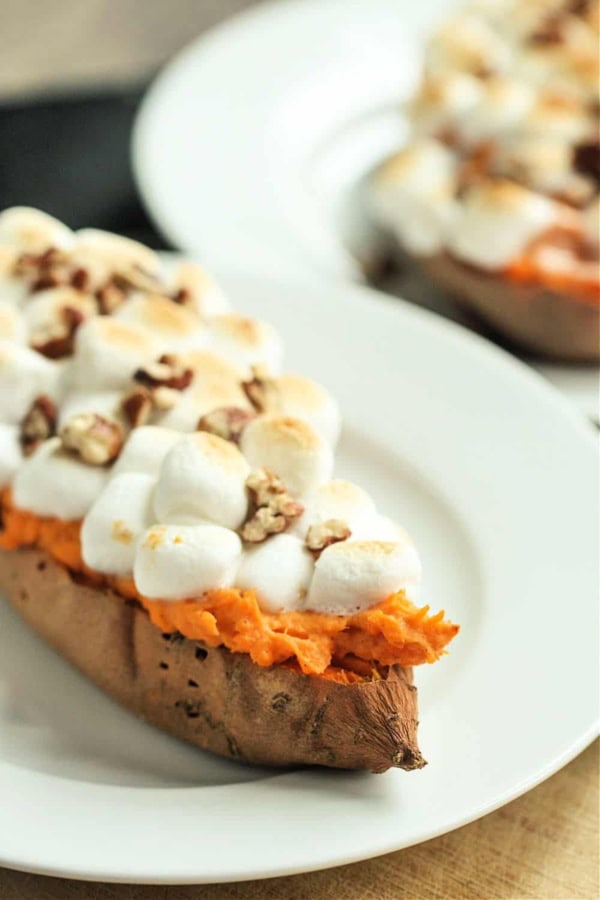 A baked sweet potato half topped with toasted marshmallows and chopped pecans is served on a white plate.