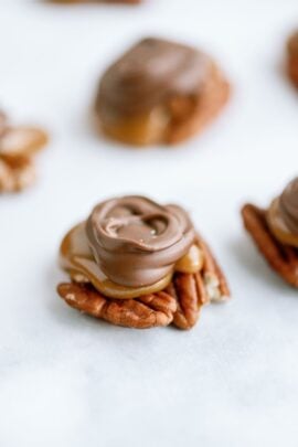 A close-up of a turtle candy featuring a pecan base, topped with caramel and covered in milk chocolate, on a white surface.