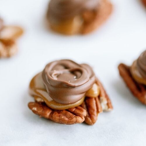 A close-up of a turtle candy featuring a pecan base, topped with caramel and covered in milk chocolate, on a white surface.