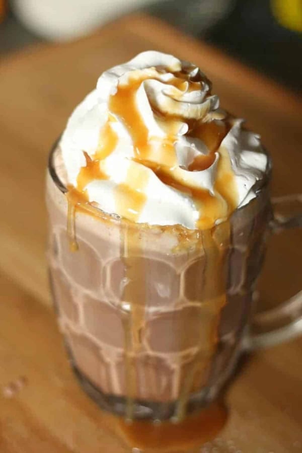 A glass mug filled with hot chocolate, topped with whipped cream, and drizzled with caramel sauce.