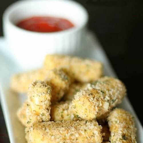 Breaded mozzarella sticks on a rectangular plate with a small white cup of red dipping sauce in the background.