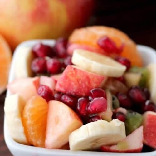 A white bowl filled with assorted fresh fruits, including banana slices, apple pieces, orange segments, kiwi, and pomegranate seeds. An apple is in the background.