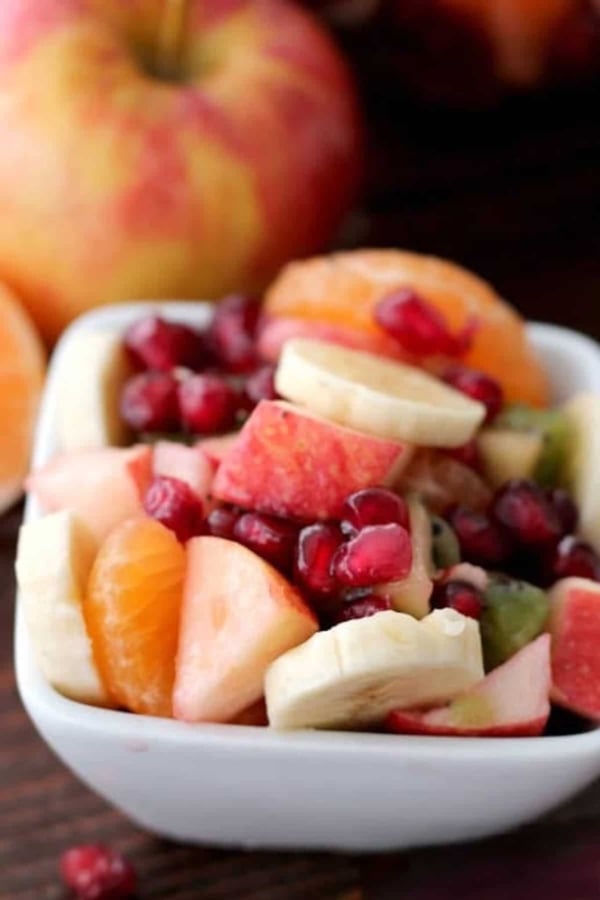 A white bowl filled with assorted fresh fruits, including banana slices, apple pieces, orange segments, kiwi, and pomegranate seeds. An apple is in the background.