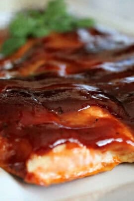 Close-up of a cooked piece of salmon fillet covered in a thick layer of barbecue sauce, with a garnish of herbs in the background.