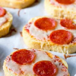 Slices of bread topped with melted cheese, tomato sauce, and pepperoni, arranged on a parchment-lined surface.