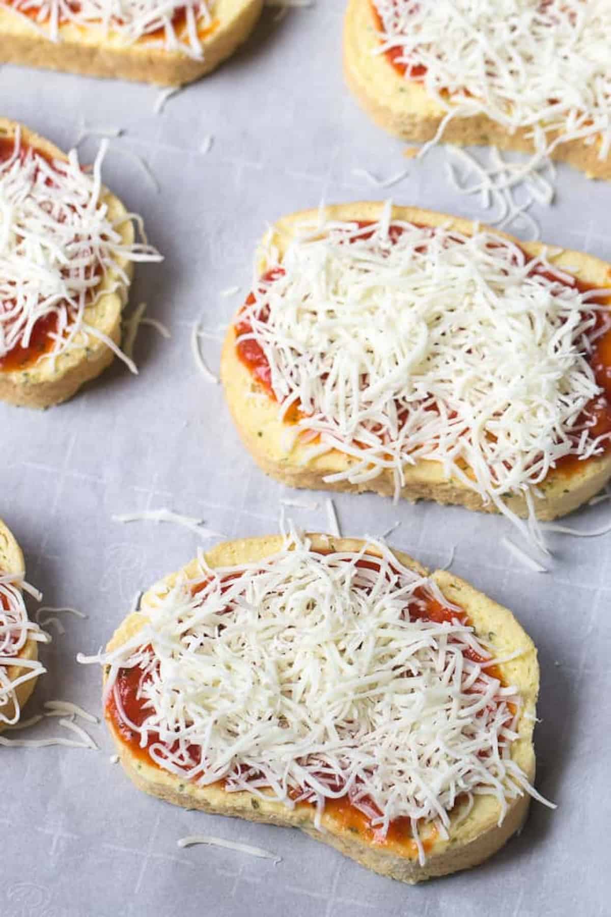 Slices of bread topped with tomato sauce and shredded mozzarella cheese are arranged on a parchment-lined surface, ready to be baked.