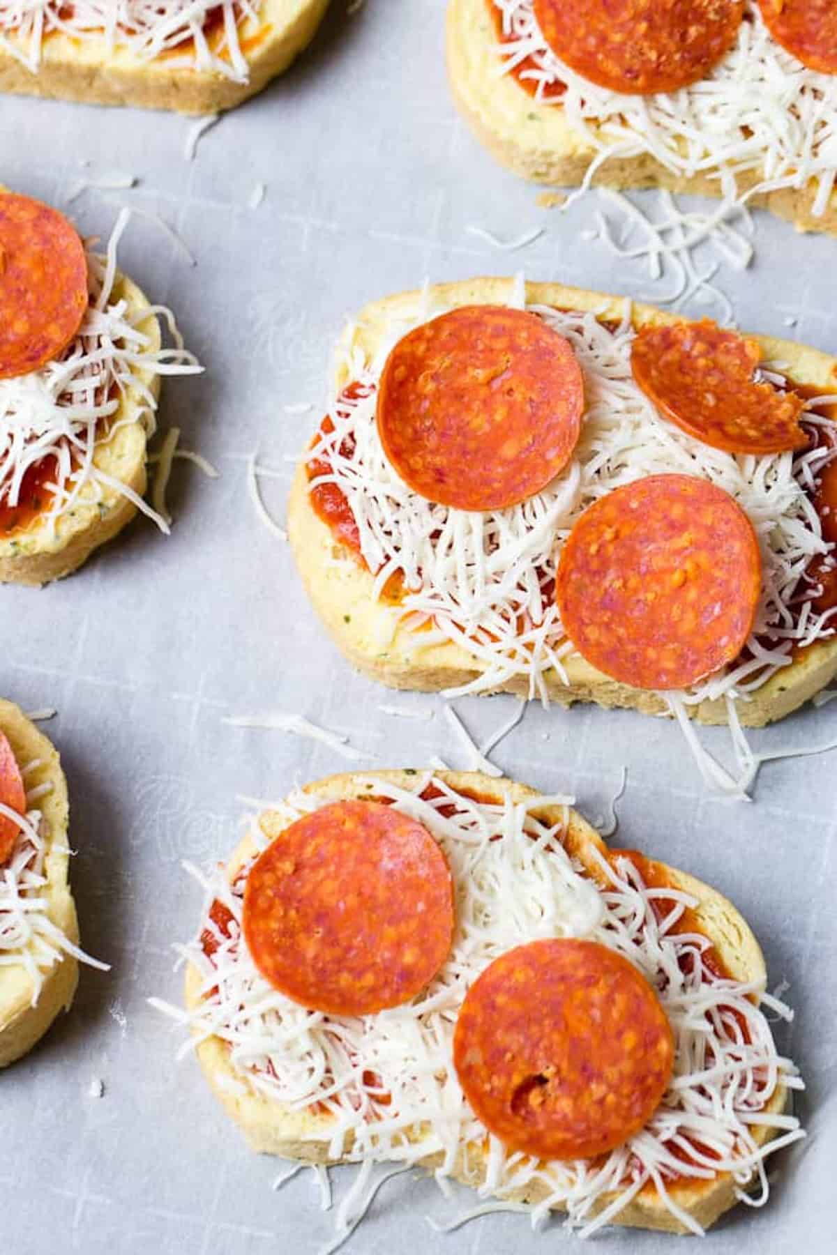 Slices of bread topped with tomato sauce, shredded cheese, and pepperoni, arranged on a parchment-lined surface, ready to be baked.