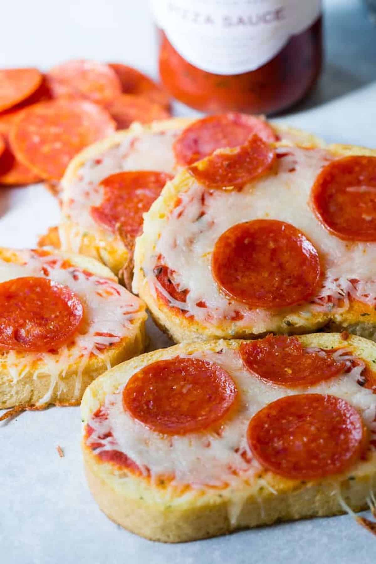 Slices of bread topped with melted cheese and pepperoni, resembling mini pizzas, with a jar of pizza sauce and pepperoni slices in the background.