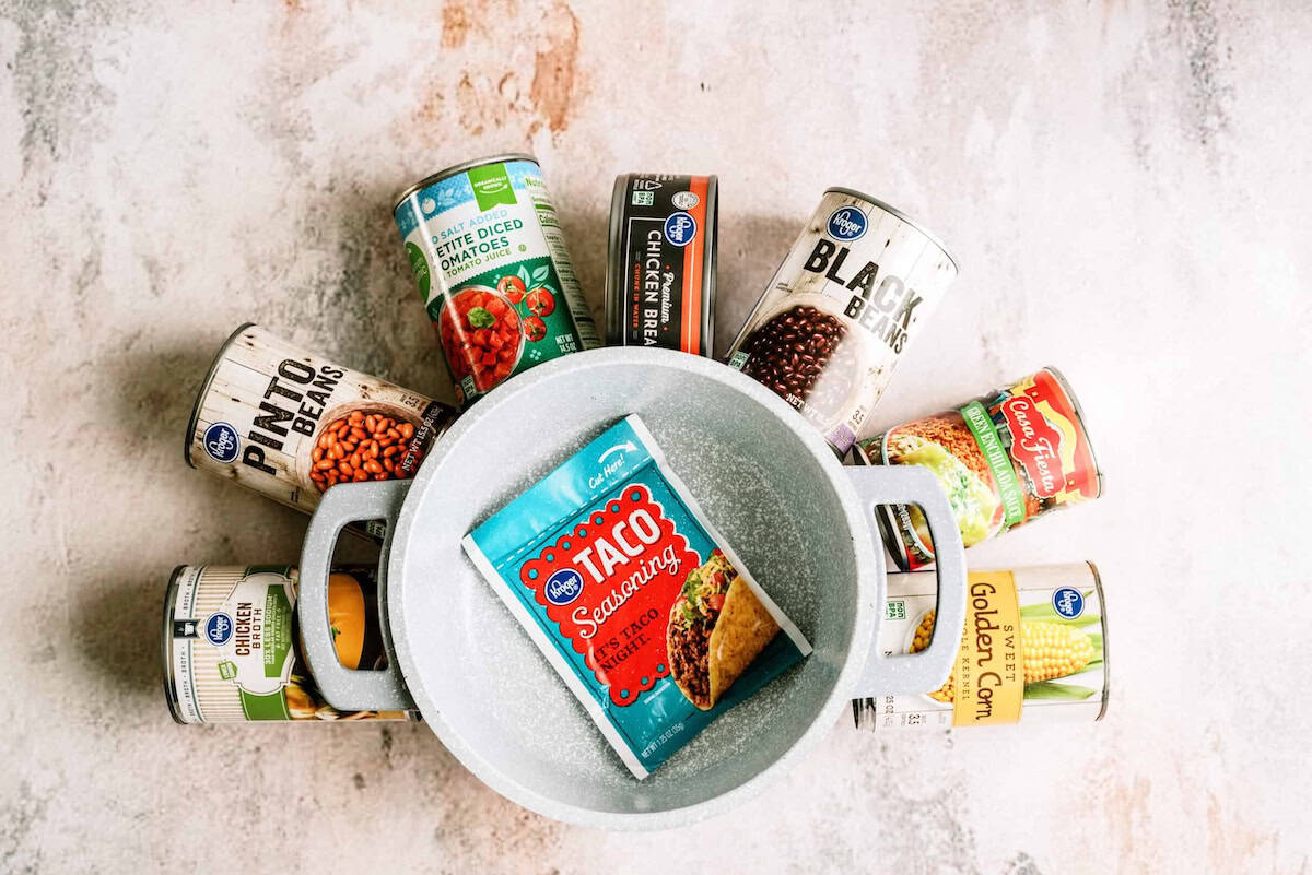 A white pot filled with a taco seasoning packet, surrounded by various canned goods including beans, tomatoes, corn, and chicken on a light surface.