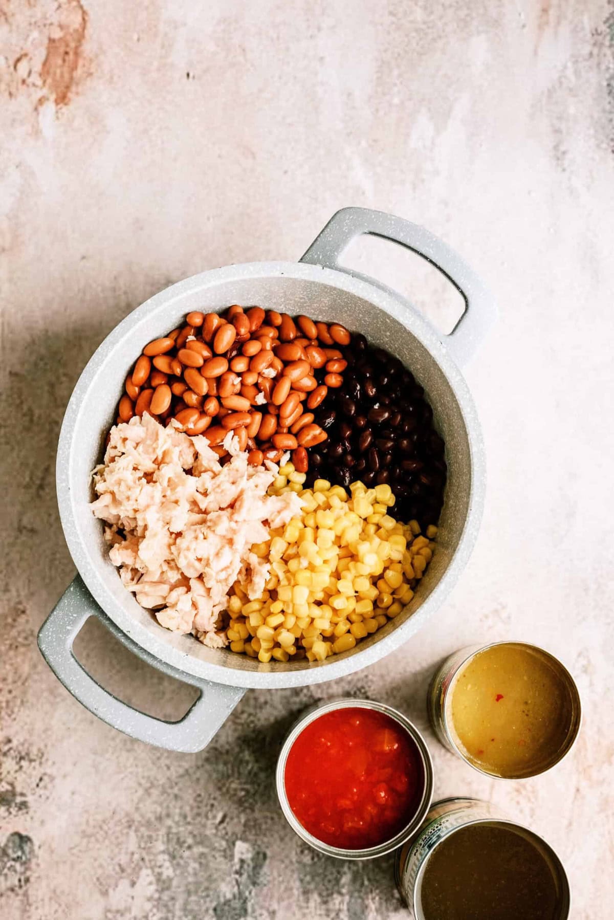 A pot containing pinto beans, black beans, corn, and shredded chicken, with three cans of broth, diced tomatoes, and green chiles nearby.