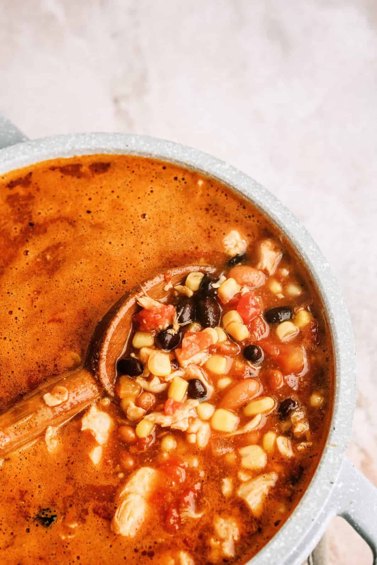 A pot of soup with a wooden ladle, showing a mixture of corn, black beans, tomatoes, and broth.