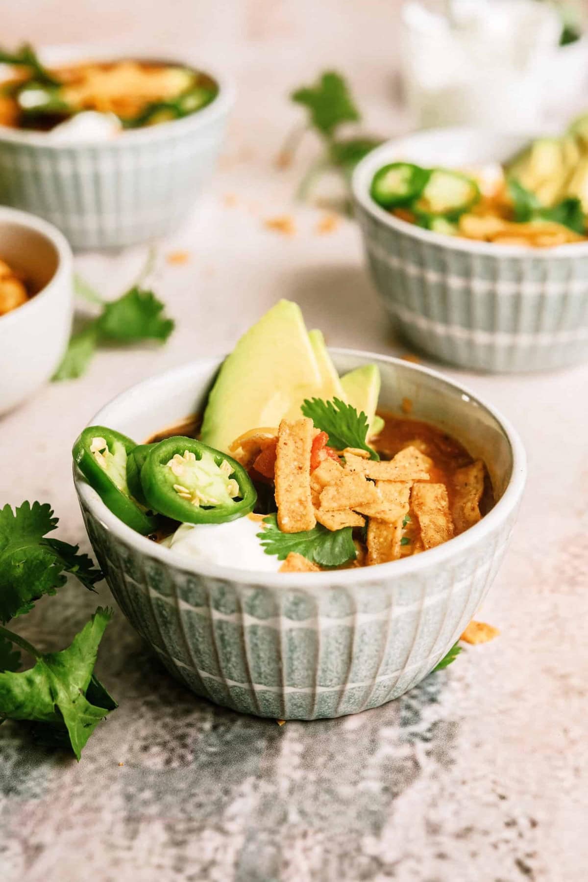 A bowl of soup topped with sliced avocado, jalapeño, tortilla strips, cilantro, and sour cream, with two similar bowls in the background.