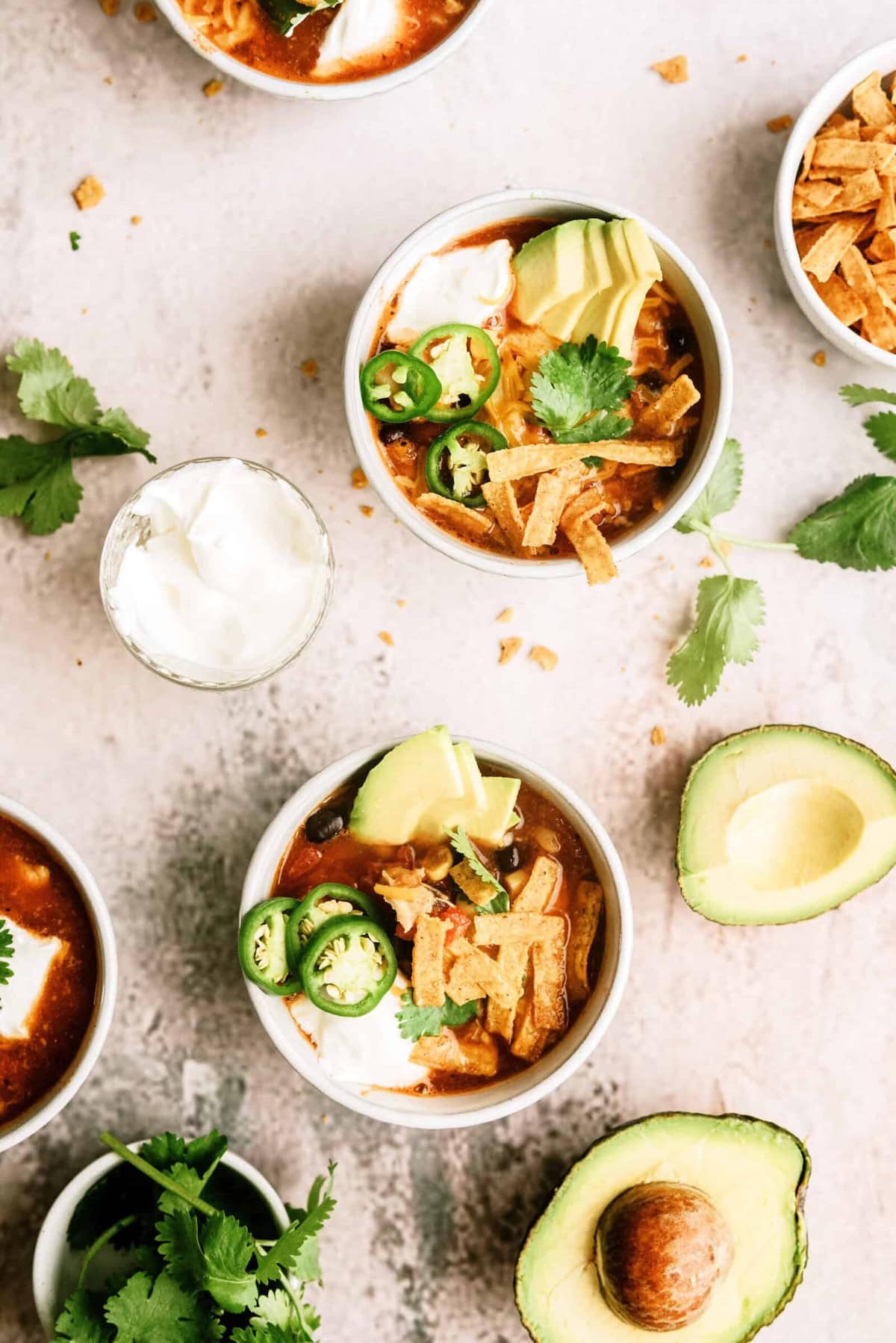 Bowls of tortilla soup topped with avocado, jalapeno, sour cream, cilantro, and tortilla strips, surrounded by fresh avocado halves and cilantro on a light surface.