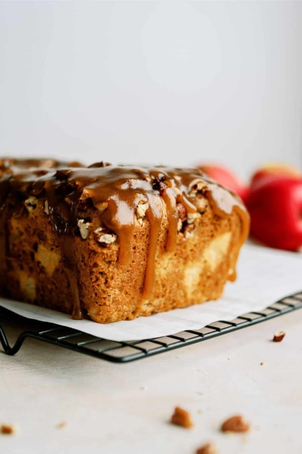 A loaf of caramel apple bread with pecans drizzled on top, placed on a cooling rack with two red apples in the background.