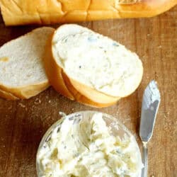A loaf of bread with two sliced pieces, one spread with herb butter. A bowl of herb butter and a butter knife are on a wooden surface.
