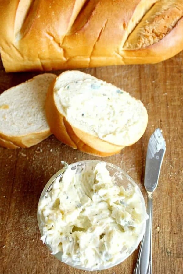 A loaf of bread with two sliced pieces, one spread with herb butter. A bowl of herb butter and a butter knife are on a wooden surface.